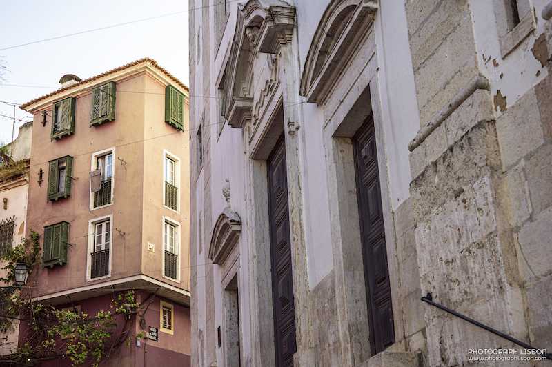 Pink facade with green shutters beside a baroque church stonework in Beco de São Miguel, Alfama, Lisbon.