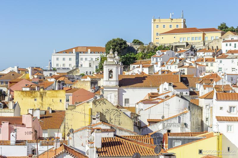 Red tiled rooftops of Alfama cascading toward a bell tower and São Jorge Castle on the hill above, Lisbon.