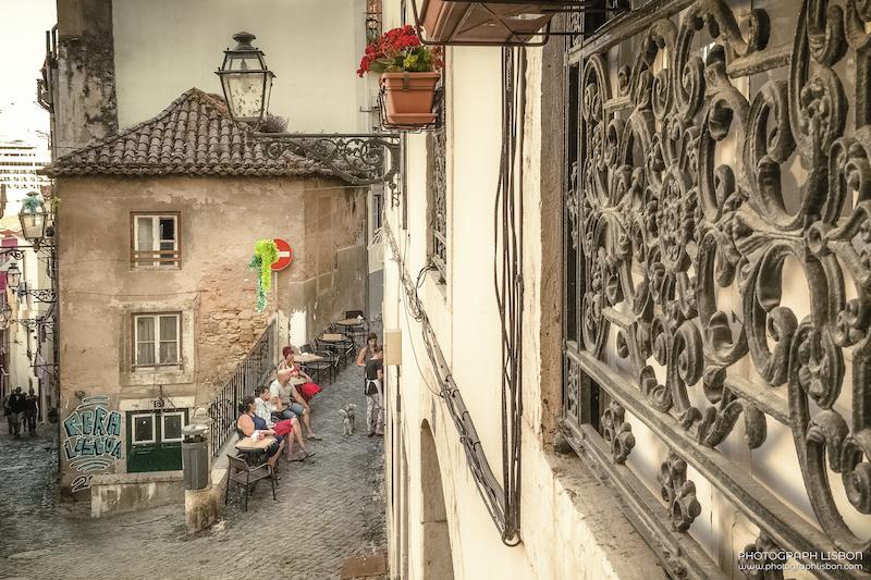 Locals at an outdoor café on cobbled stairs with a wrought-iron street lamp and ornate balcony, Alfama, Lisbon.