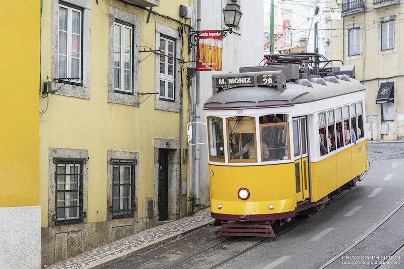 Tram 28 on its route through a narrow street lined with yellow facades in Lisbon's old town.