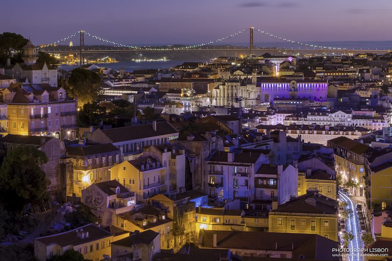 Panoramic view of Lisbon at blue hour with the 25 de Abril Bridge lit in the distance.