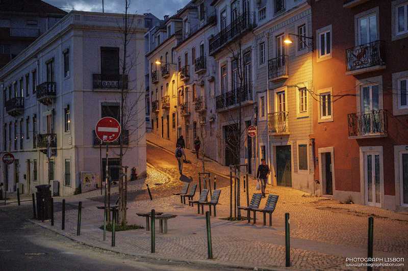 Cobbled Lisbon street at dusk lit by warm street lamps, with pedestrians walking up the slope.