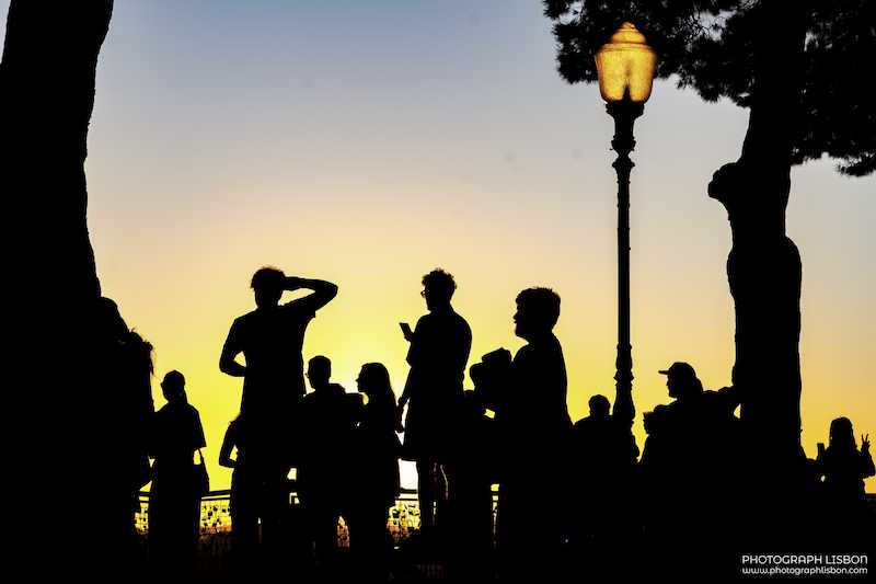 Silhouettes of visitors and a wrought-iron street lamp against a golden sunset sky at a Lisbon viewpoint.