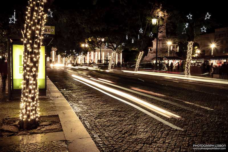 Long-exposure traffic trails along Rossio at night, framed by trees wrapped in Christmas lights, Lisbon.